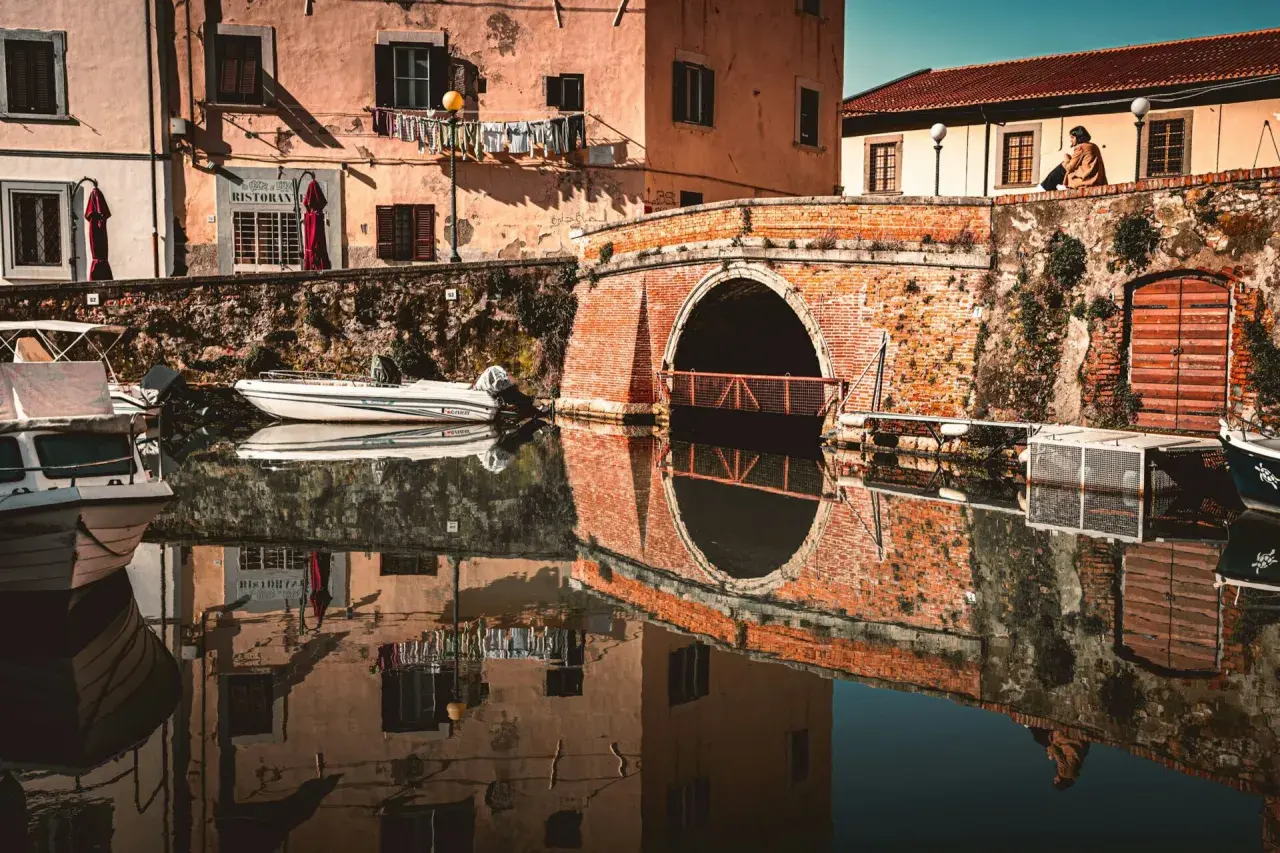 Un ponte in mattoni rossi attraversa un canale nel quartiere Venezia di Livorno. Barche ormeggiate riflettono sulle acque calme, mentre panni stesi aggiungono un tocco di vita quotidiana.