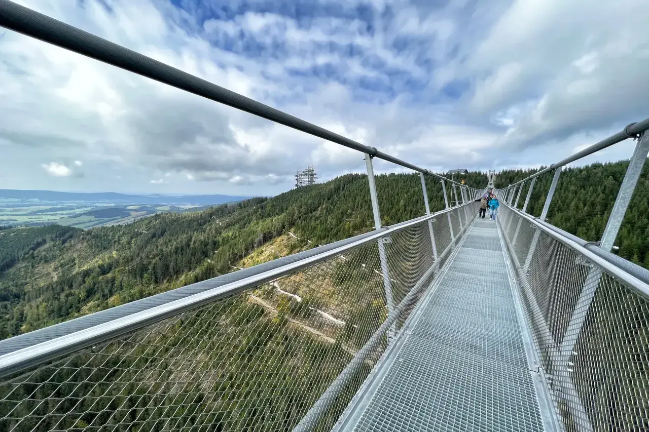 Himmelbrücke in Dolní Morava mit Blick auf bewaldete Hügel und eine Fernsehturm-ähnliche Struktur.