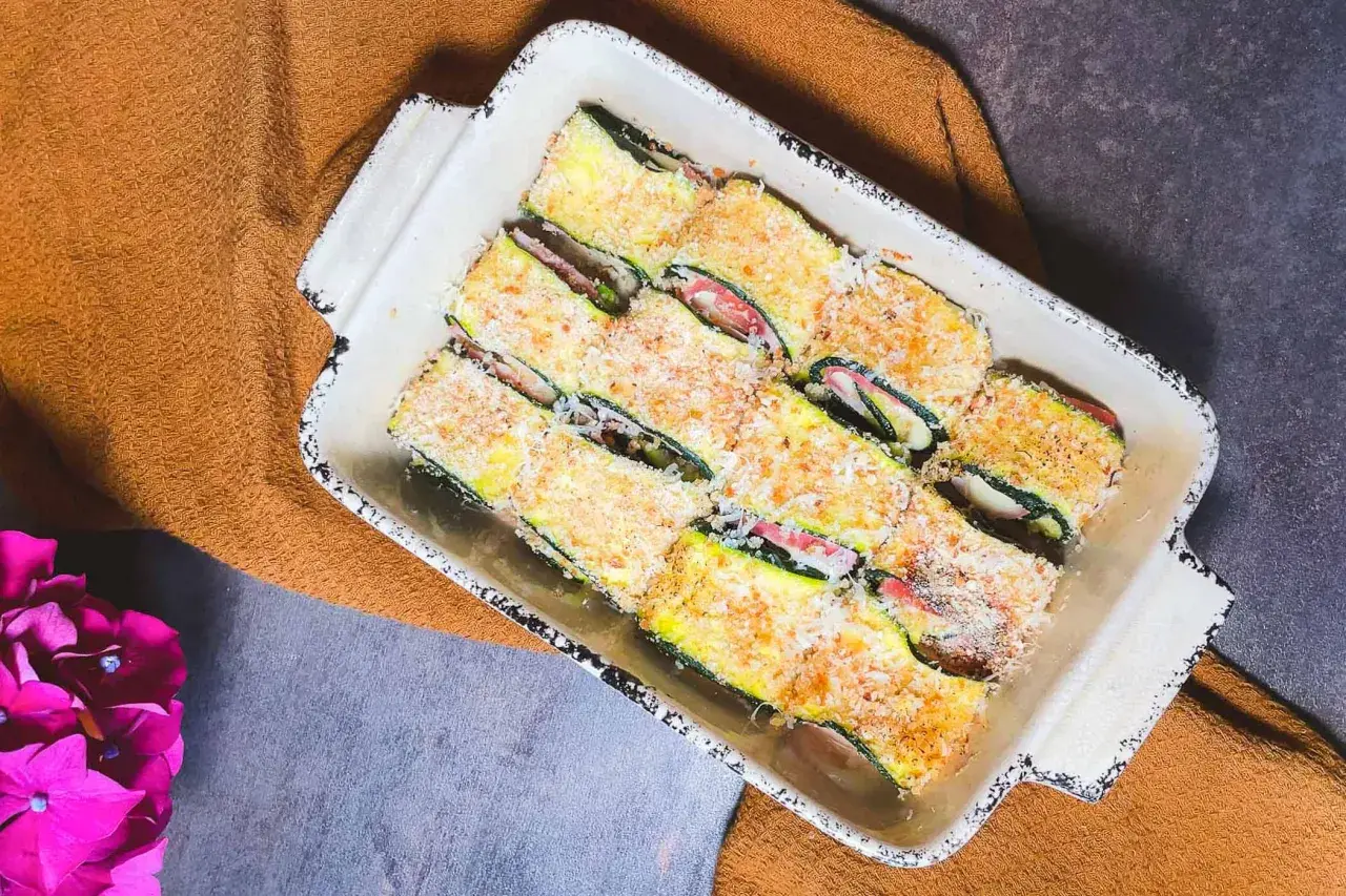 Involtini di zucchine pronti per essere cotti, ripieni e impanati, in una teglia bianca su un tovagliolo arancione.