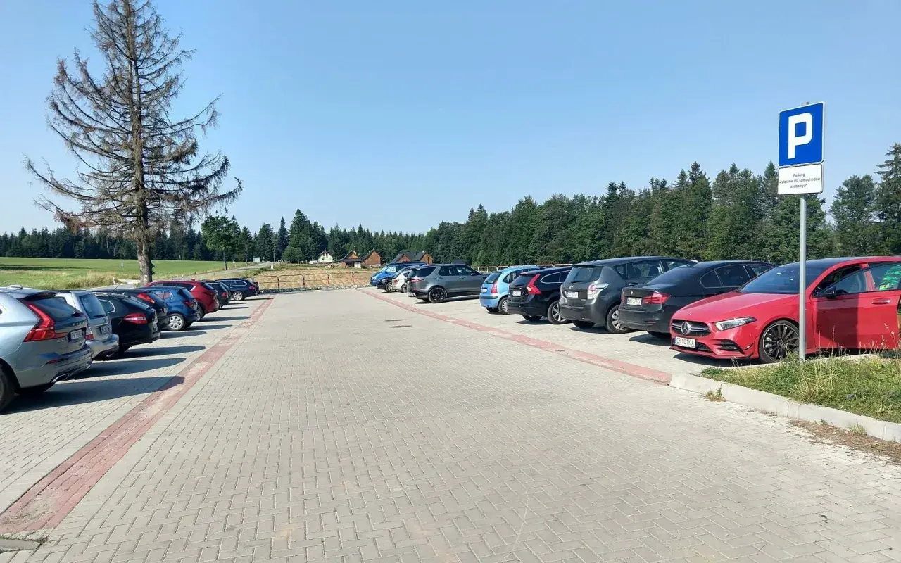 Parking przy górze Jagodnej. Samochody zaparkowane na utwardzonym placu, w tle las i domki.