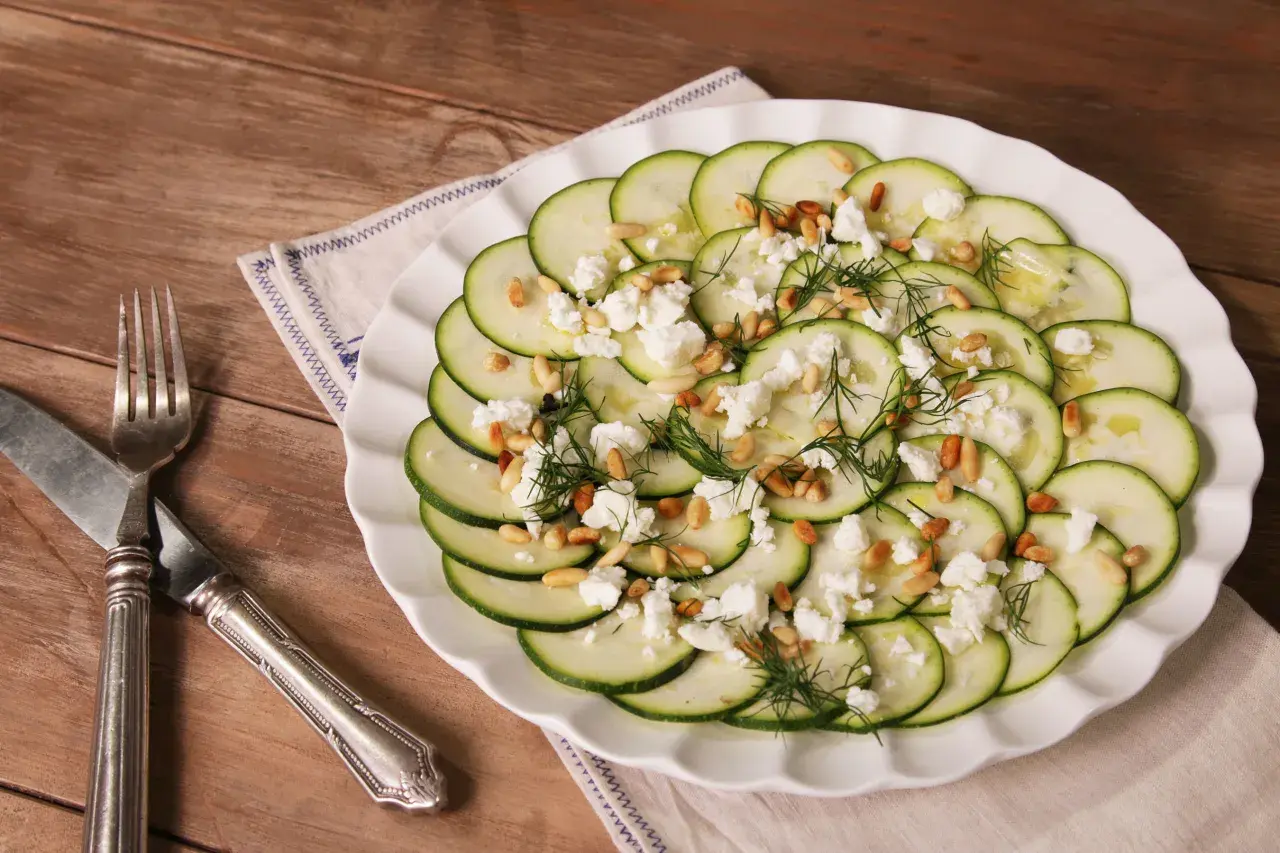 Delicioso carpaccio de calabacín con queso feta desmenuzado, piñones tostados y eneldo fresco, servido en un plato blanco sobre una mesa de madera.
