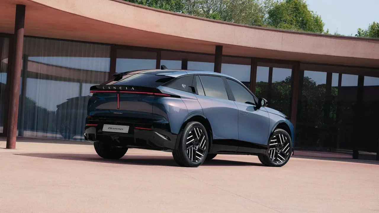 Lancia Gamma 2026, un'elegante berlina blu metallizzato con fari posteriori a LED, parcheggiata davanti a un edificio moderno.