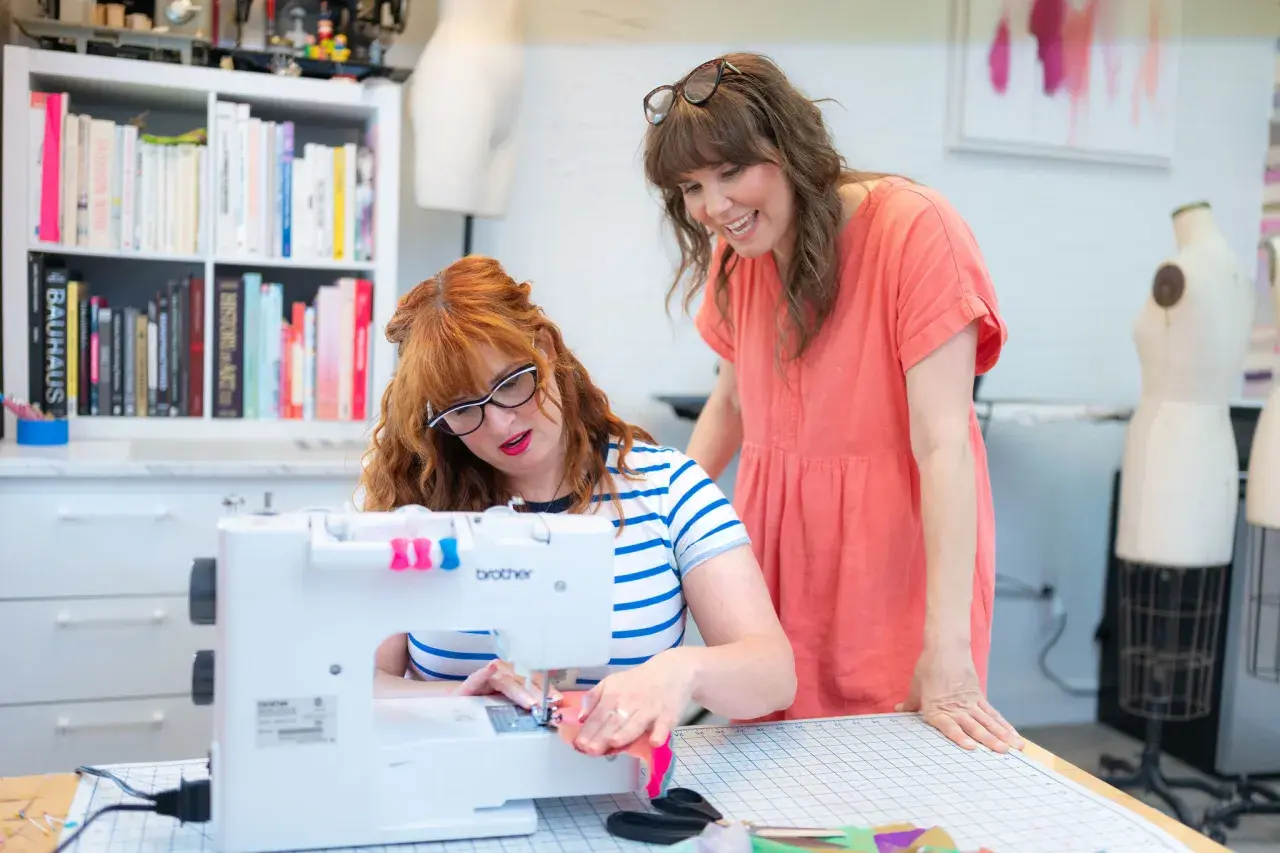 Two women are focused on sewing seams on a white Brother sewing machine in a bright studio.