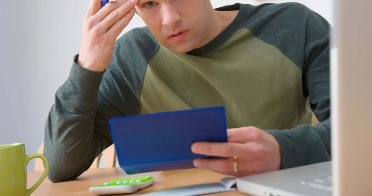 Homme pensif, calculatrice, tasse verte, ordinateur portable. Il se demande combien de temps garder chéquier.