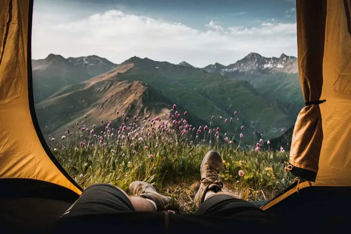 Vista desde una tienda de campaña: piernas con botas de montaña, flores silvestres y montañas verdes bajo un cielo nublado. Disfrutando del acampar con tienda de campaña.