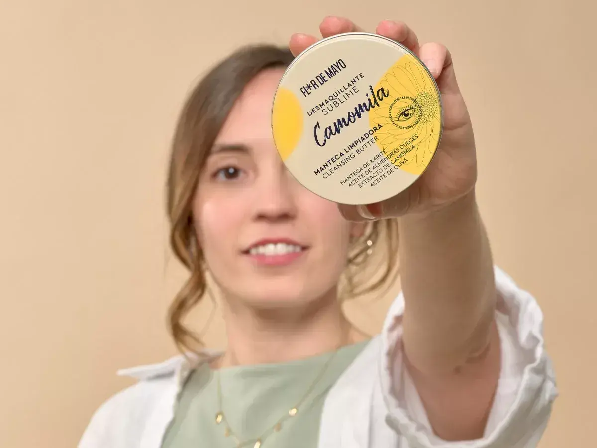 Mujer sonriente sostiene un desmaquillante en crema, "Camomila", que es un sebo limpiador con manteca de karité.
