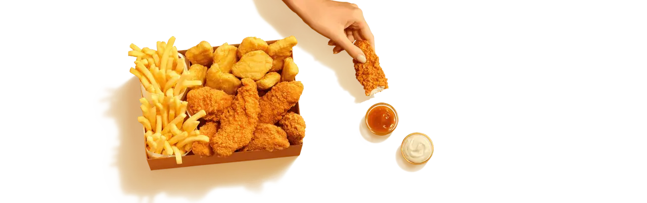 Pudełko McDonald's z kurczakiem i frytkami. Ręka zanurza kawałek kurczaka w sosie. Sprawdź kcal!