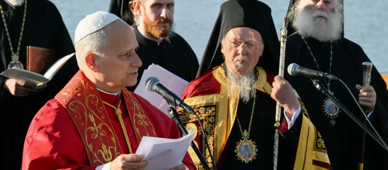Zwei Geistliche, einer im roten Gewand, der andere mit Bart und Mitra, stehen vor Mikrofonen. Der Mann mit Mitra ist ein Patriarch.