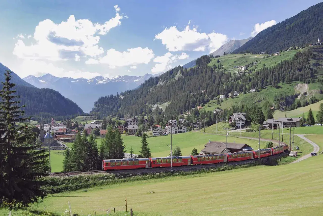 Ein roter Zug fährt durch eine malerische Alpenlandschaft. Ein perfekter tagesausflug mit der bahn, vorbei an grünen Wiesen, Wäldern und einem Dorf mit schneebedeckten Bergen im Hintergrund.