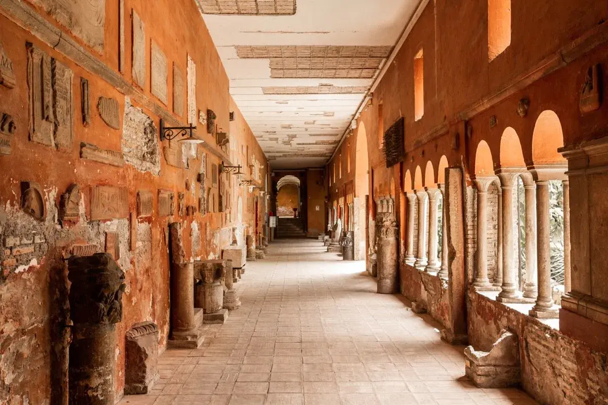 Corridoio con reperti antichi, un'idea di cosa vedere a Trastevere. Colonne, iscrizioni e frammenti decorano le pareti arancioni.