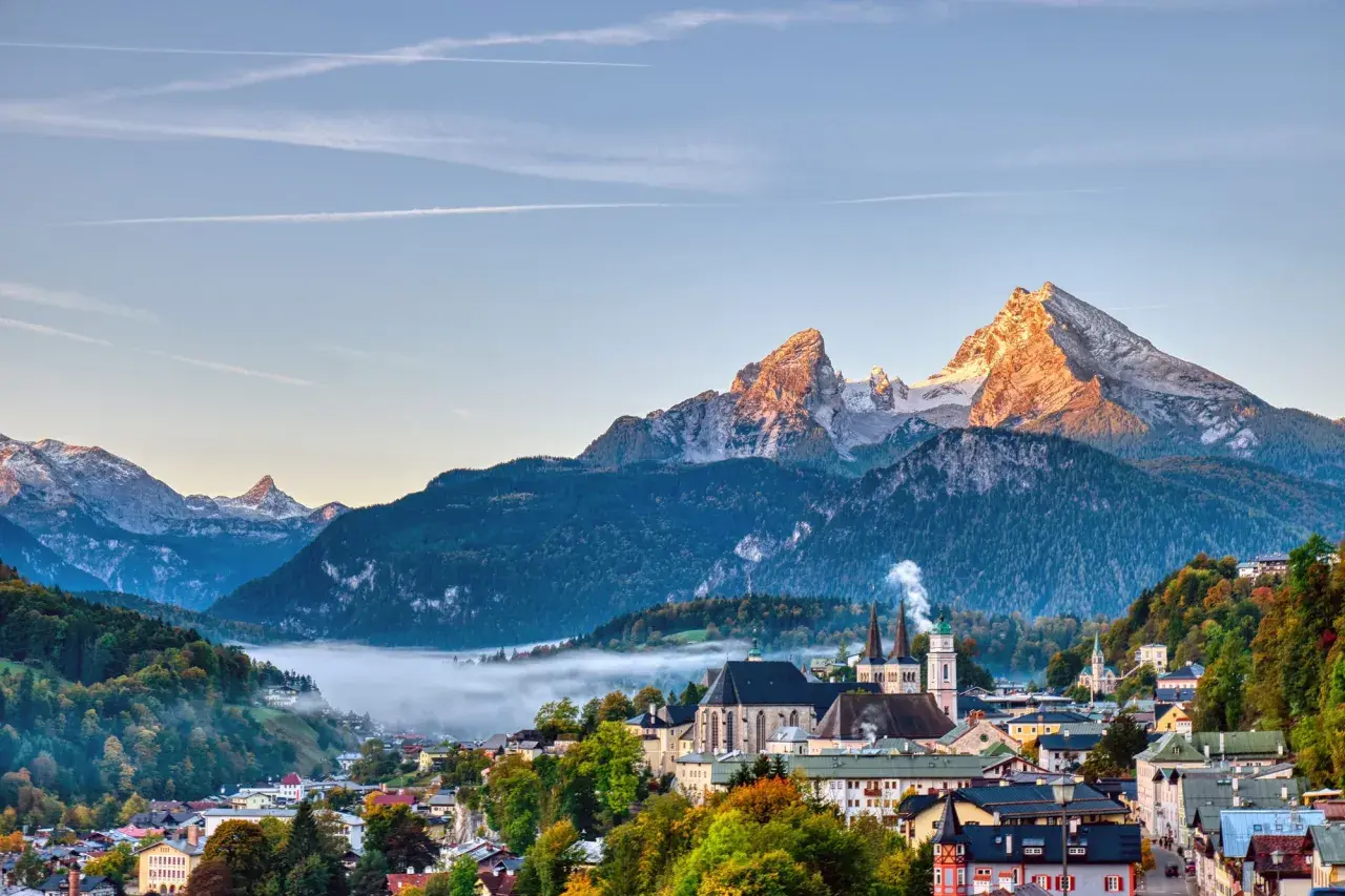 Morgensonne beleuchtet den spitzen Gipfel des kleinen Watzmanns über einem nebelverhangenen Tal und einer malerischen Stadt mit Kirchtürmen.
