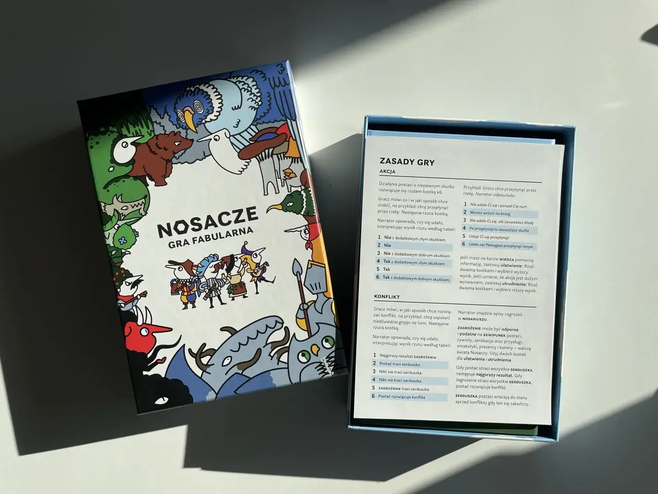 Pudełko gry fabularnej "Nosacze" z kolorowymi ilustracjami i otwartą instrukcją.