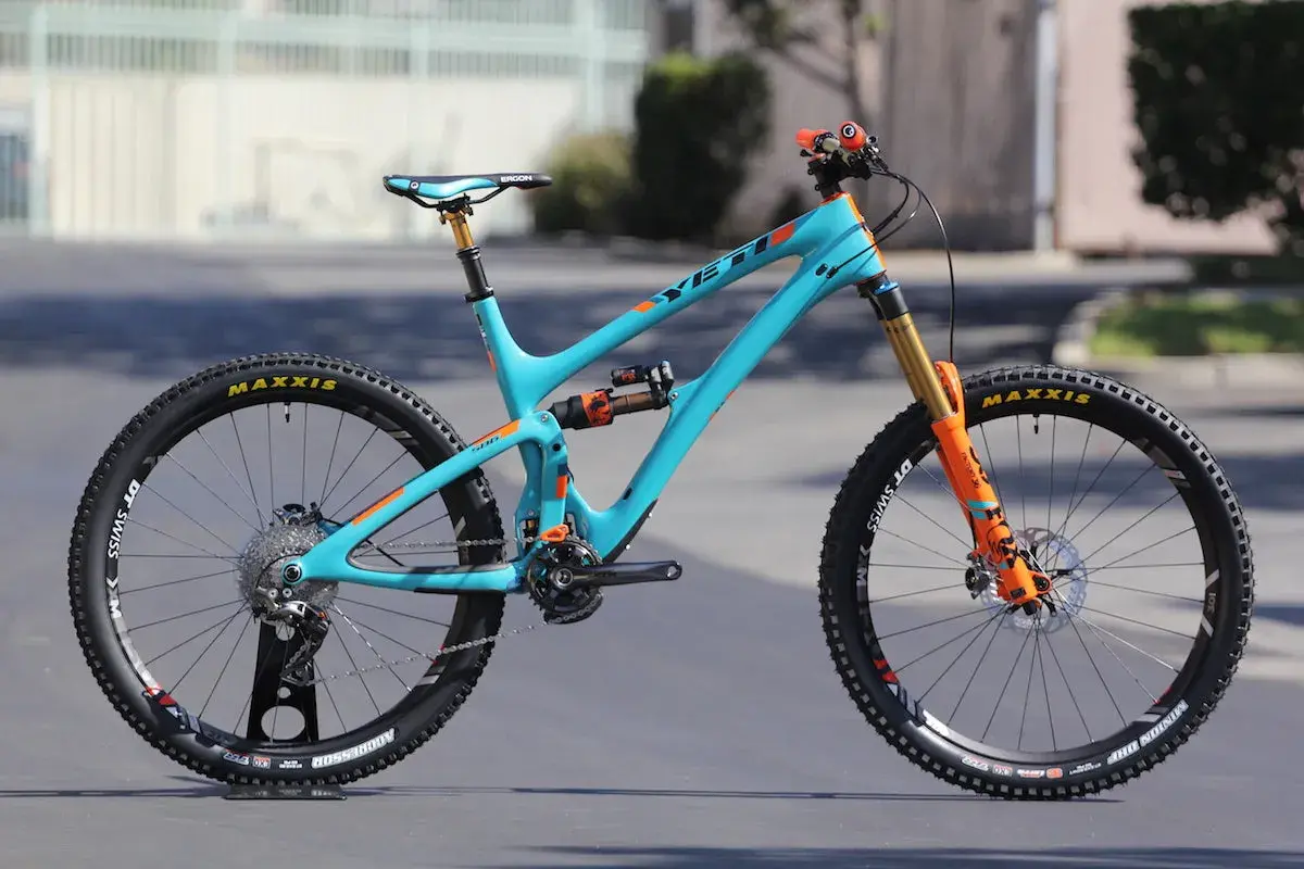 A turquoise Yeti SB6c mountain bike with orange accents, featuring Maxxis tires and Fox suspension, ready for any trail.