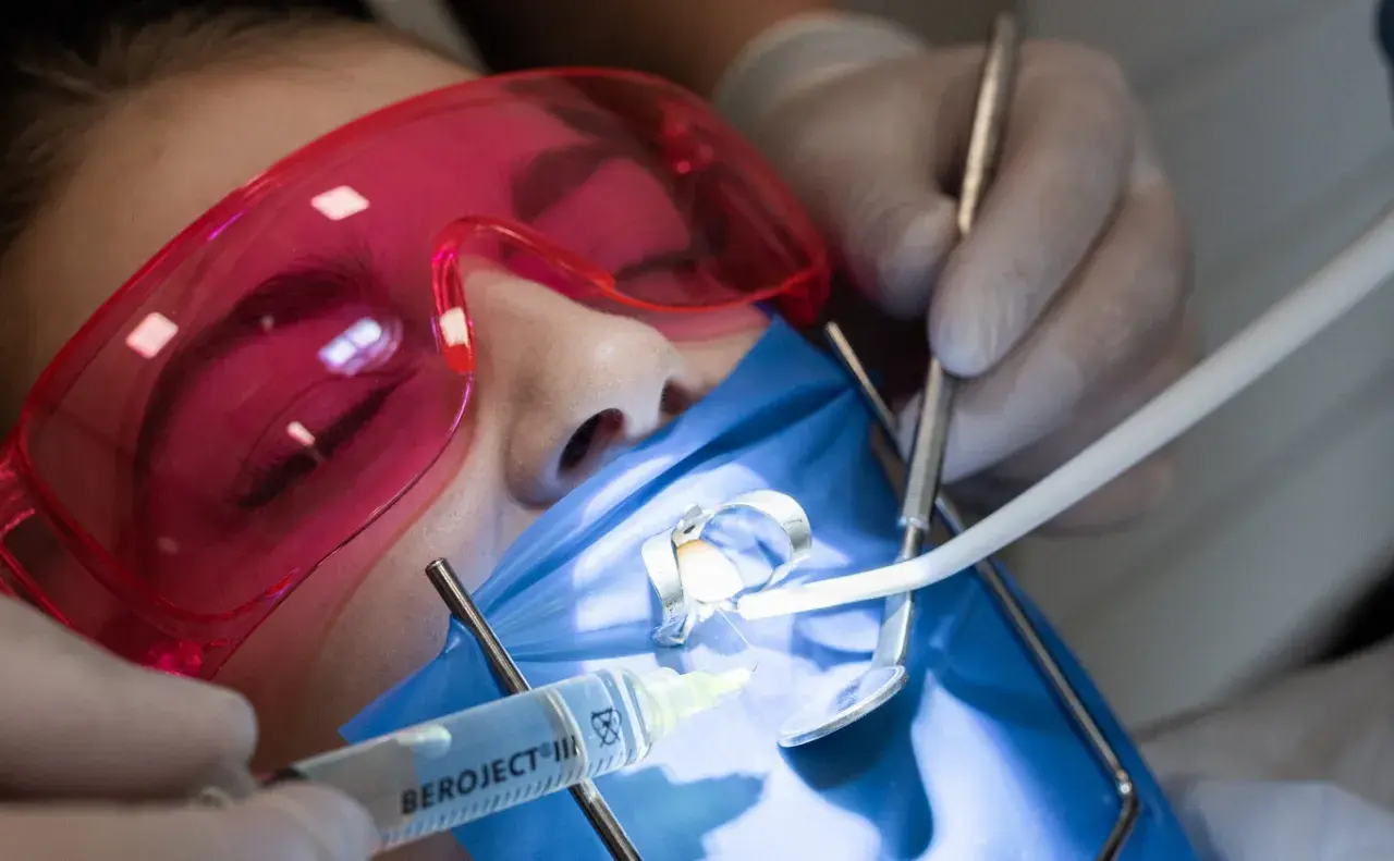 Precyzyjne leczenie kanałowe pod mikroskopem. Dentysta w rękawiczkach używa narzędzi i strzykawki. Pacjent ma założone różowe okulary ochronne.