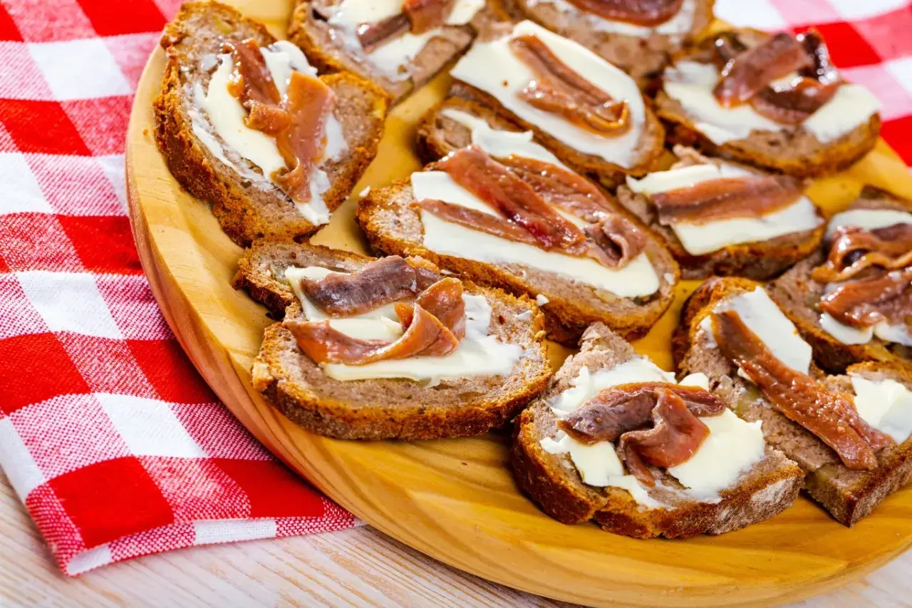 Kromki chleba z masłem i filecikami anchois ułożone na drewnianej desce, obok czerwono-biała serwetka.