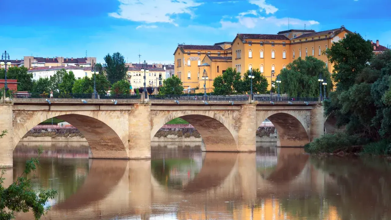 Puente de piedra sobre el río Ebro, reflejándose en sus aguas. Edificios amarillos y verdes árboles componen el paisaje de **que ver en Logroño**.