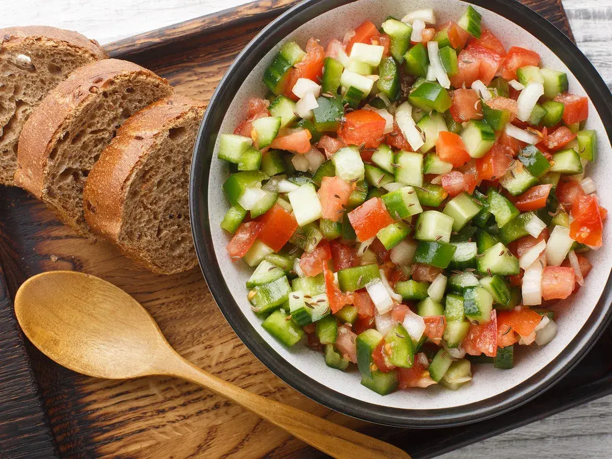 Ensalada pipirrana fresca y colorida, con tomate, pepino, pimiento y cebolla, servida junto a rebanadas de pan integral y una cuchara de madera.