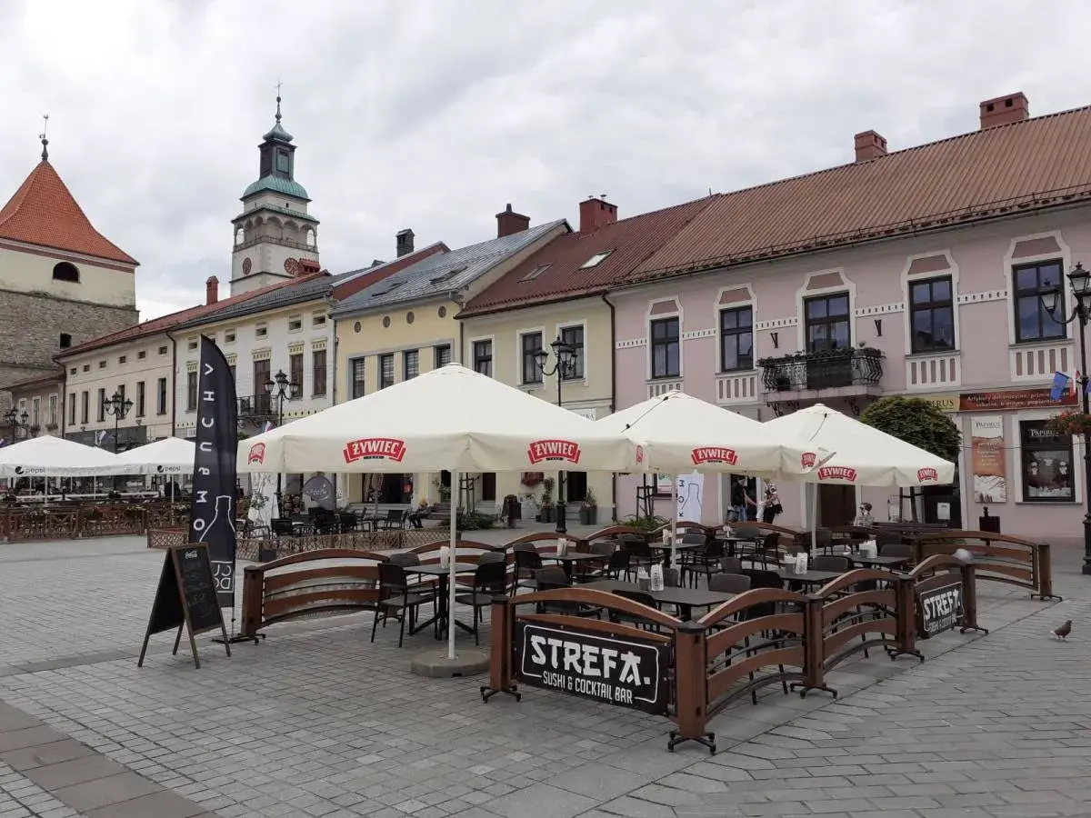 Letni ogródek kawiarniany na rynku w Żywcu. Parasole z logo Żywiec, stoliki i ławki. W tle kamienice i wieża ratuszowa.
