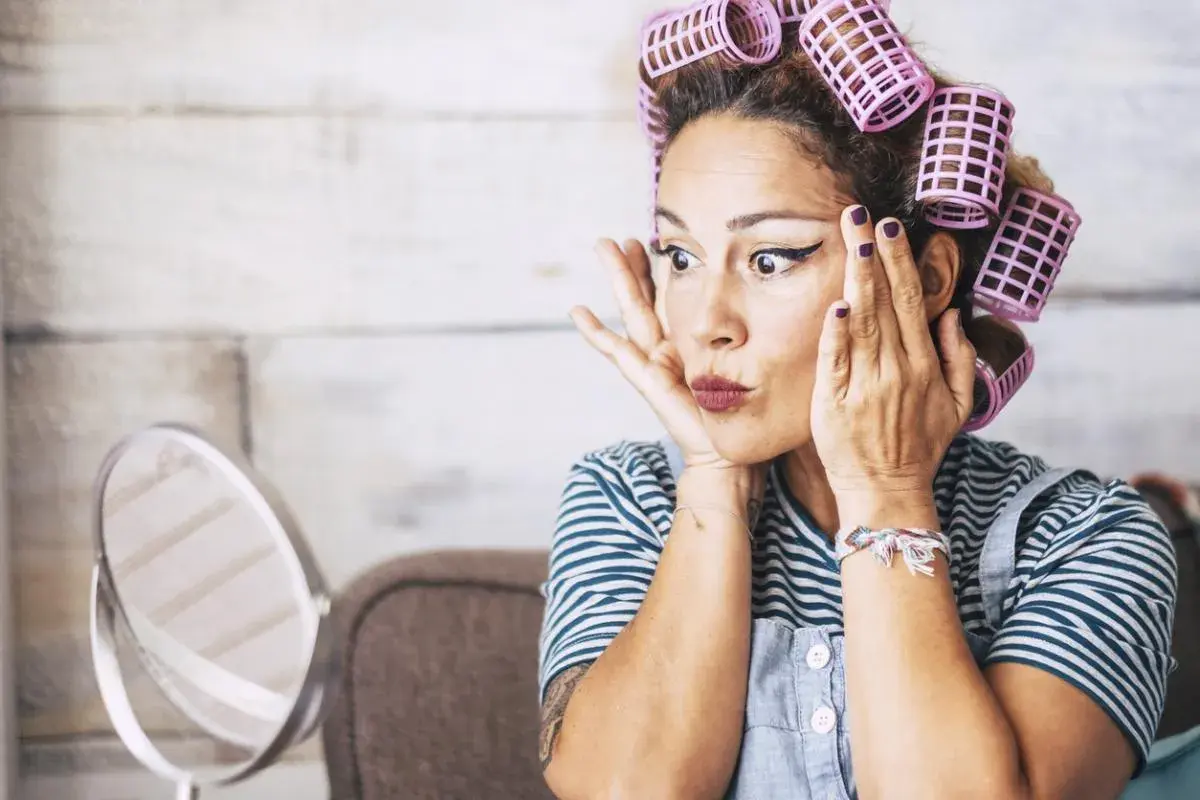 Mujer con rulos en el pelo, delineado de ojos y labios pintados, practicando cómo rizar el pelo frente a un espejo.