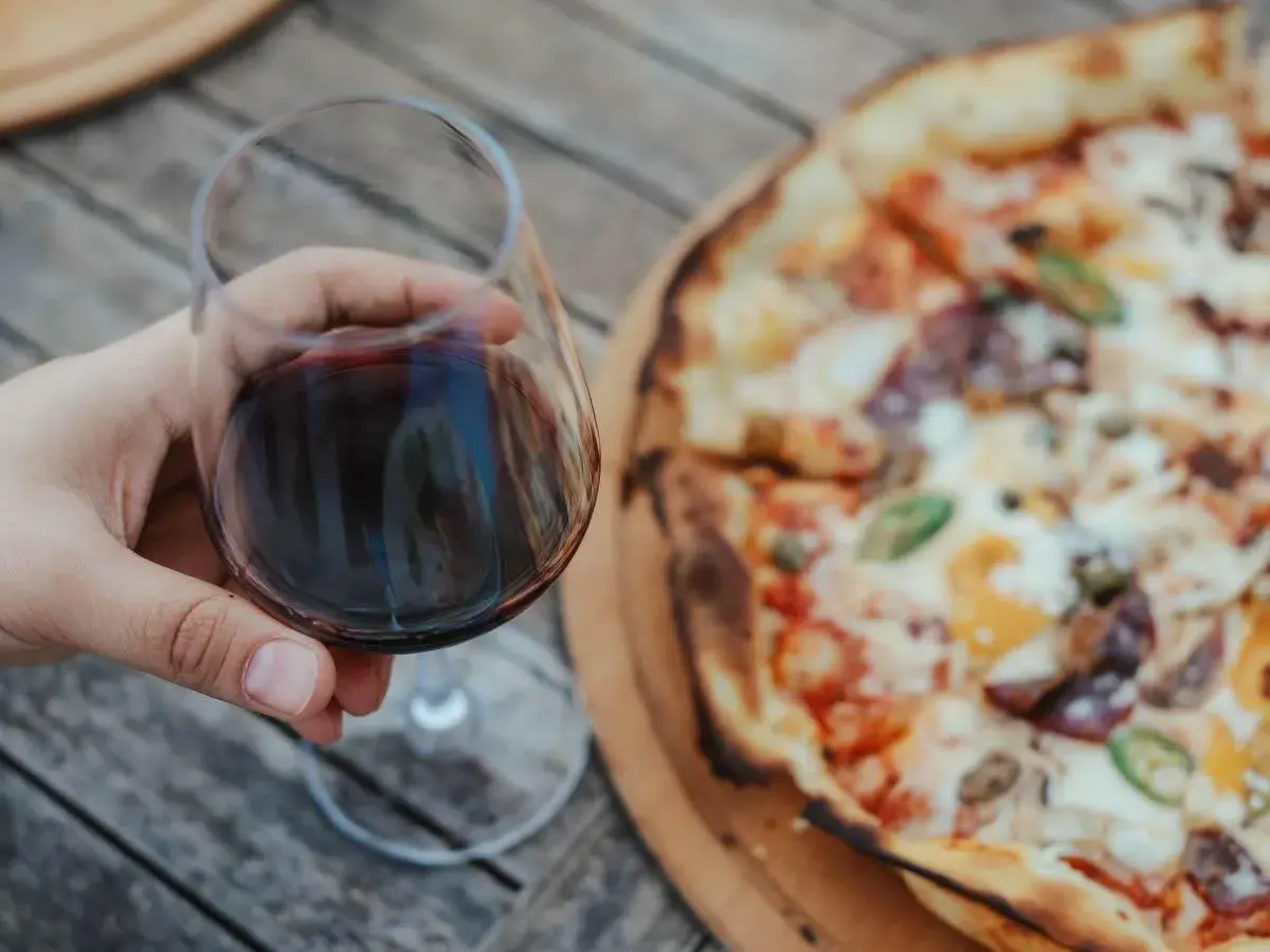 Un bicchiere di vino rosso in mano, accanto a una gustosa pizza. Un abbinamento perfetto per una serata all'aperto.