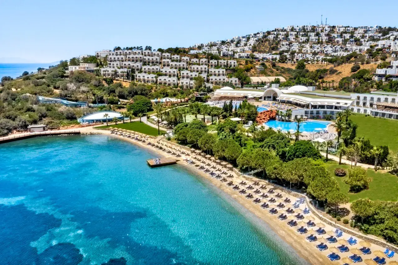 Widok na hotel Yasmin Bodrum Resort z plażą, basenami i białymi budynkami na wzgórzu. Idealne miejsce na wakacje, jak sugerują yasmin bodrum resort opinie.