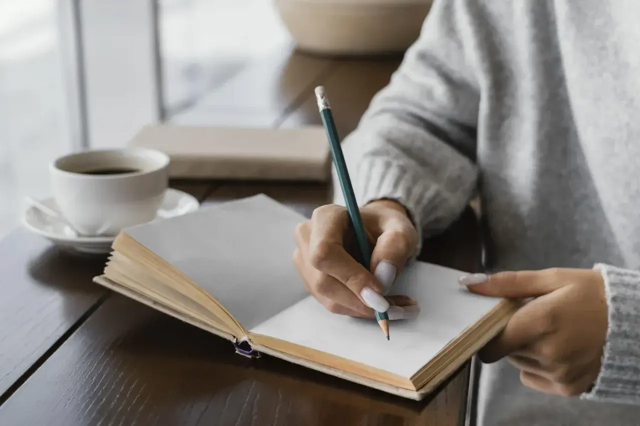 Mano escribiendo en un diario, un ritual de autocuidado. El café humeante acompaña este momento de introspección, una forma de escribir un diario como terapia.