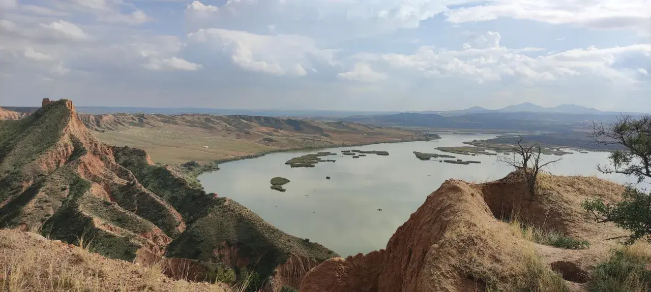 Paisaje de barrancos ocres y un lago sereno, ideal para una ruta de senderismo.