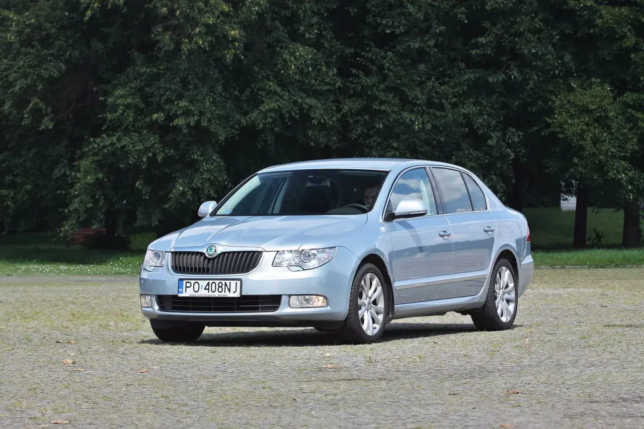 Jasnoniebieska Skoda Superb 2 z nowymi żarówkami, zaparkowana na placu przed drzewami.