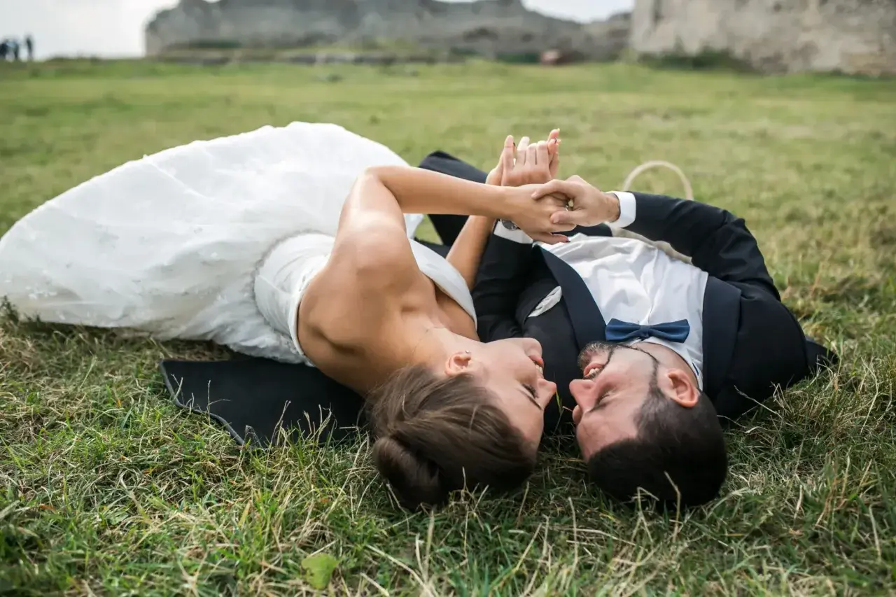 Para młoda leży na trawie podczas sesji plenerowej ślubnej, trzymając się za ręce. W tle ruiny zamku.
