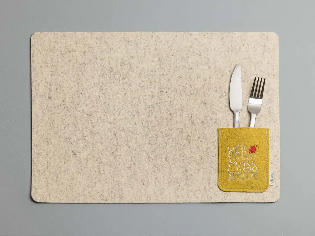 Beiges Platzset aus Filz mit gelber Tasche und der Aufschrift "Wer kleckert muss spülen!". Messer und Gabel stecken darin.