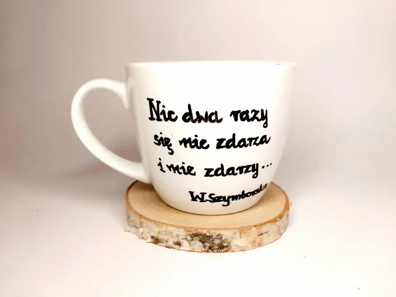 Biały kubek z cytatem Wisławy Szymborskiej "Nic dwa razy się nie zdarza..." na drewnianym podkładzie.