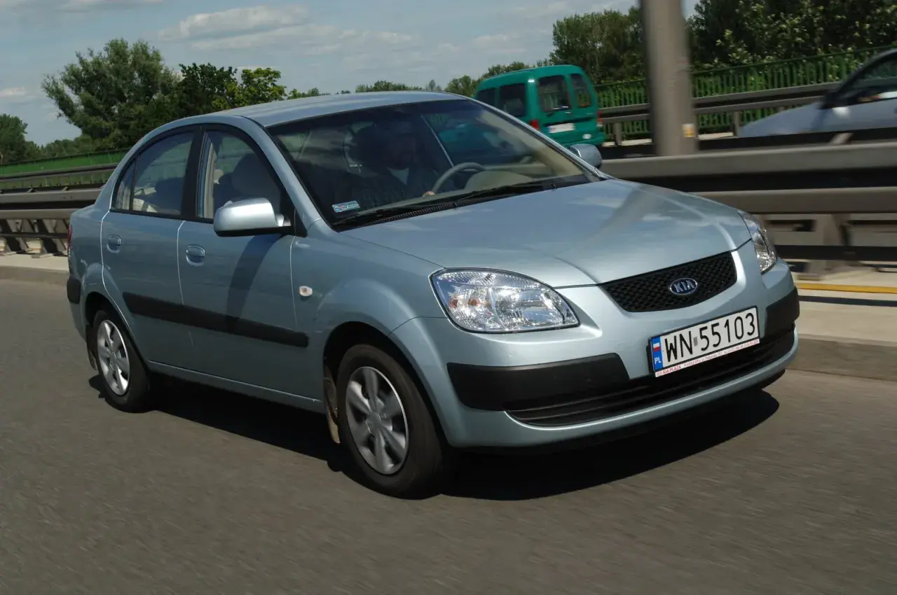 Błękitne światła Kia Rio na drodze. Kierowca w okularach prowadzi auto. W tle zielony van i drzewa.