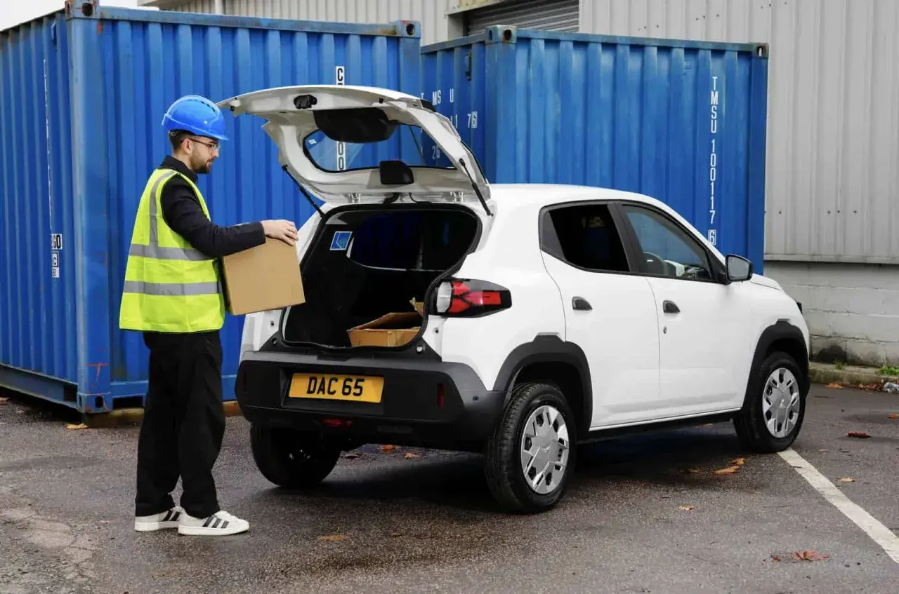 Pracownik w kamizelce odblaskowej wkłada paczkę do bagażnika białego Dacia Spring Cargo, gotowego do dostawy.
