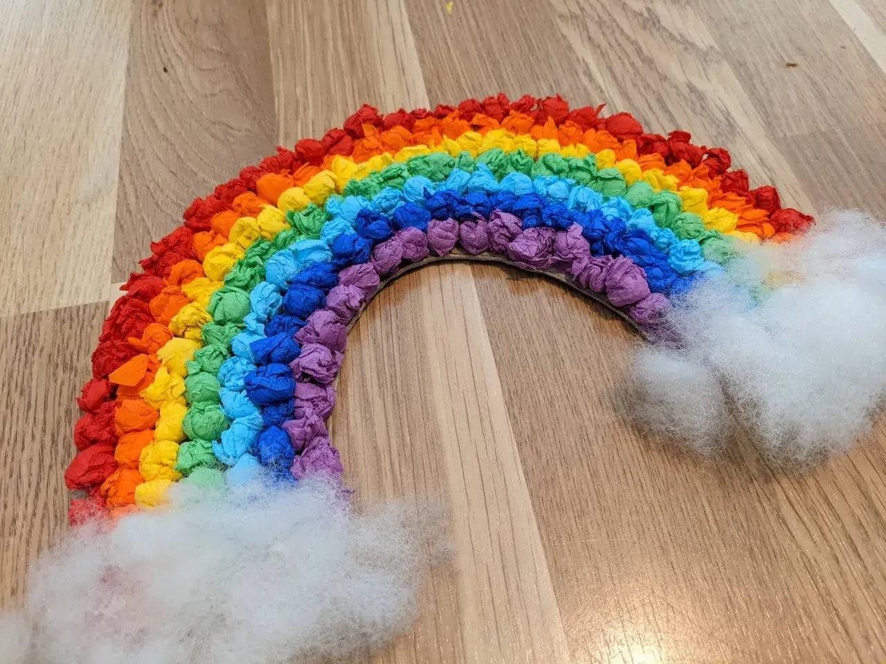 Ein bunter Regenbogen, gebastelt aus zerknülltem Krepppapier in allen Farben, mit flauschigen Wolken aus Watte.