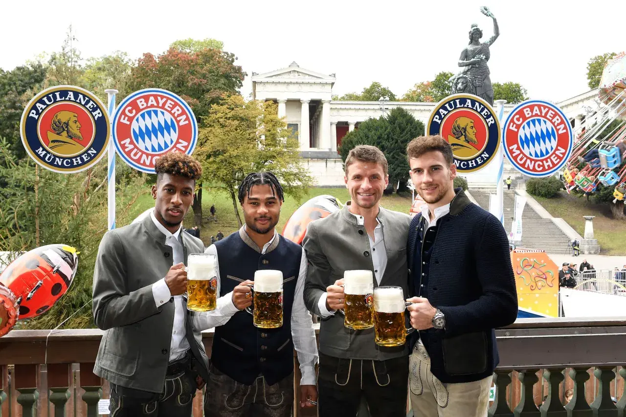 Vier FC Bayern Spieler in Tracht mit Bierkrügen, vor Paulaner- und FC Bayern-Logos. Ein Fest für alle Fans bei Veranstaltungen in Bayern.