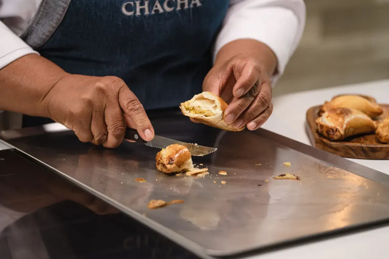 Manos cortan una empanada dorada, revelando su relleno. Otra masa empanada espera en una tabla.