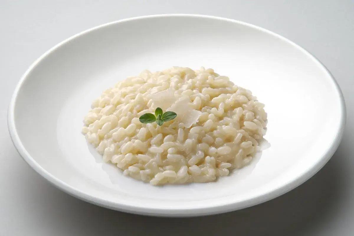 Un cremoso risotto in cagnone, guarnito con parmigiano e una fogliolina verde, servito in un piatto bianco.