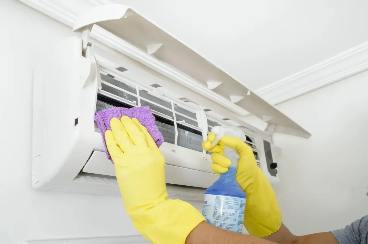 Man con guantes amarillos limpia un aire acondicionado con un paño morado y spray. Aprende como desatascar un aire acondicionado.