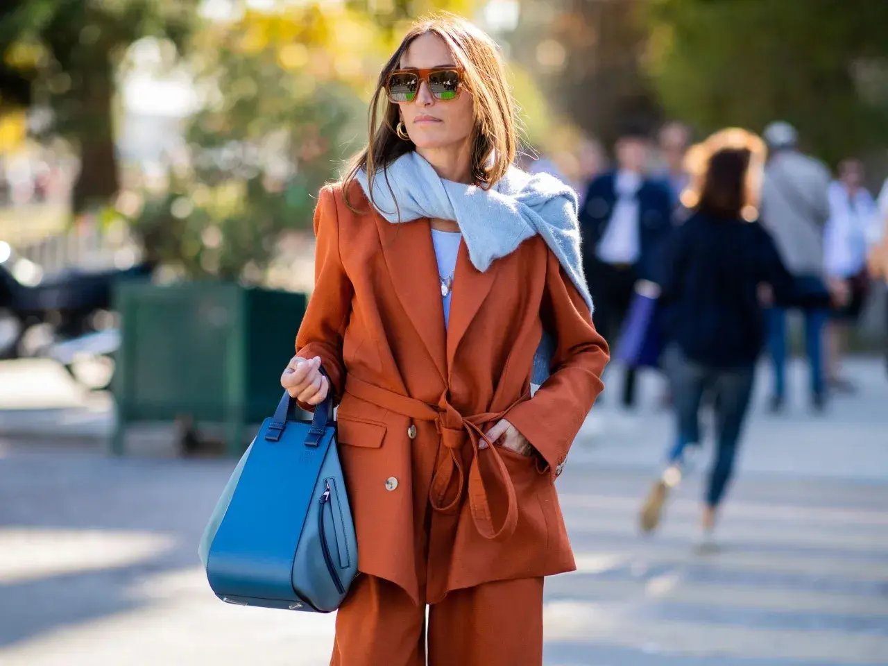 Stilvolle Frau mit braunem Anzug und hellblauem Schal. Ihre Tasche kombiniert blau und braun, ein modischer Hingucker.