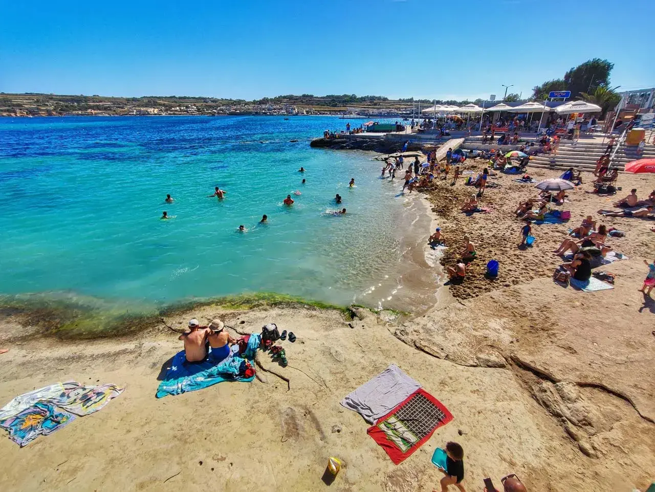 Słoneczna plaża z turkusową wodą, na której kąpią się ludzie, a na piasku odpoczywają plażowicze.