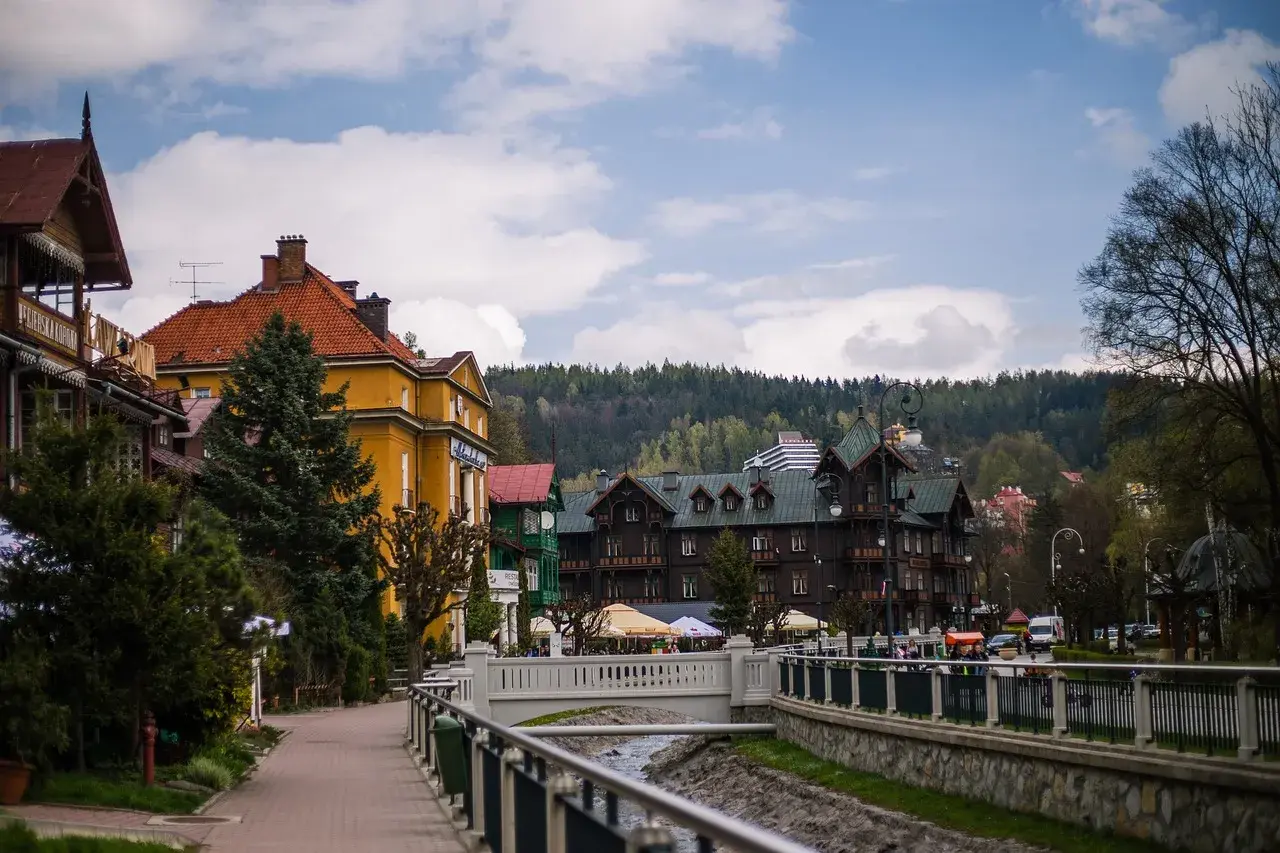 Urokliwa promenada w Krynicy-Zdroju, z widokiem na zabytkowe wille i górskie zbocza. W oddali widać budynek z napisem "Polonus", przypominający o podróży z Warszawy Zachodniej.