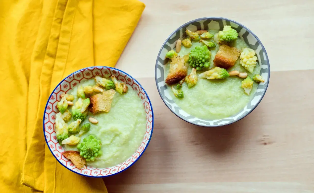 Zwei Schalen cremiger Gemüsesuppe, garniert mit Romanesco, Croutons und Pinienkernen. Ein köstliches Romanesco-Rezept für kalte Tage.