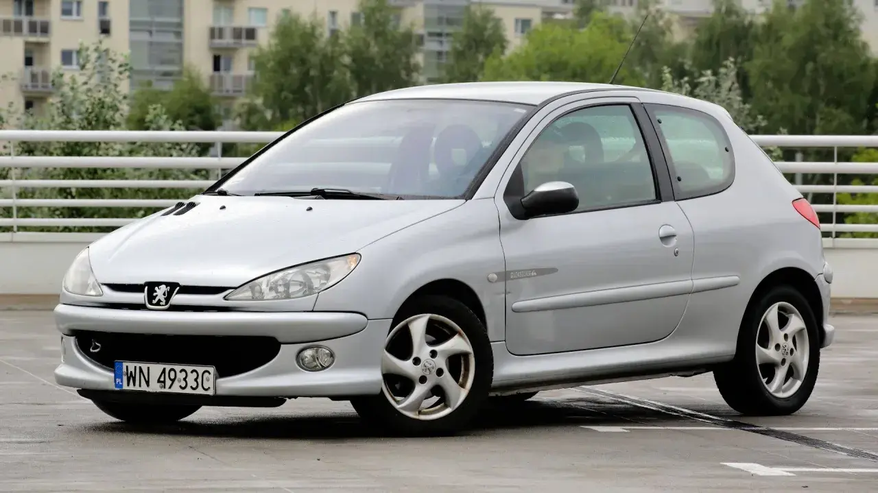 Srebrny Peugeot 206 z widocznymi przednimi żarówkami, zaparkowany na mokrym parkingu.
