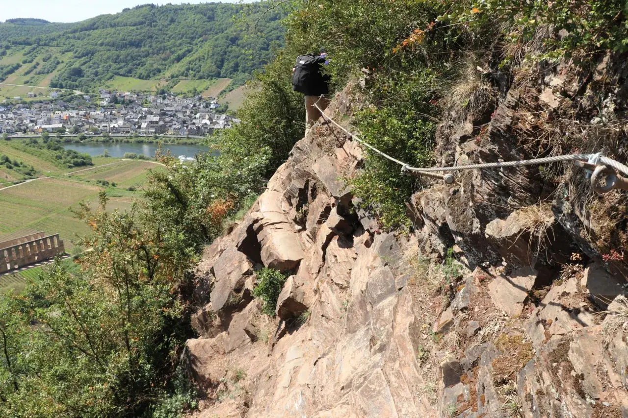 Wanderer sichert sich mit Seil am Felssteig über der Mosel. Blick auf Weinberge und Dorf.