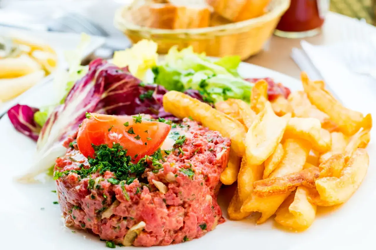 Un délicieux tartare de boeuf, garni d'une tranche de tomate et de persil, servi avec des frites croustillantes et une salade fraîche.