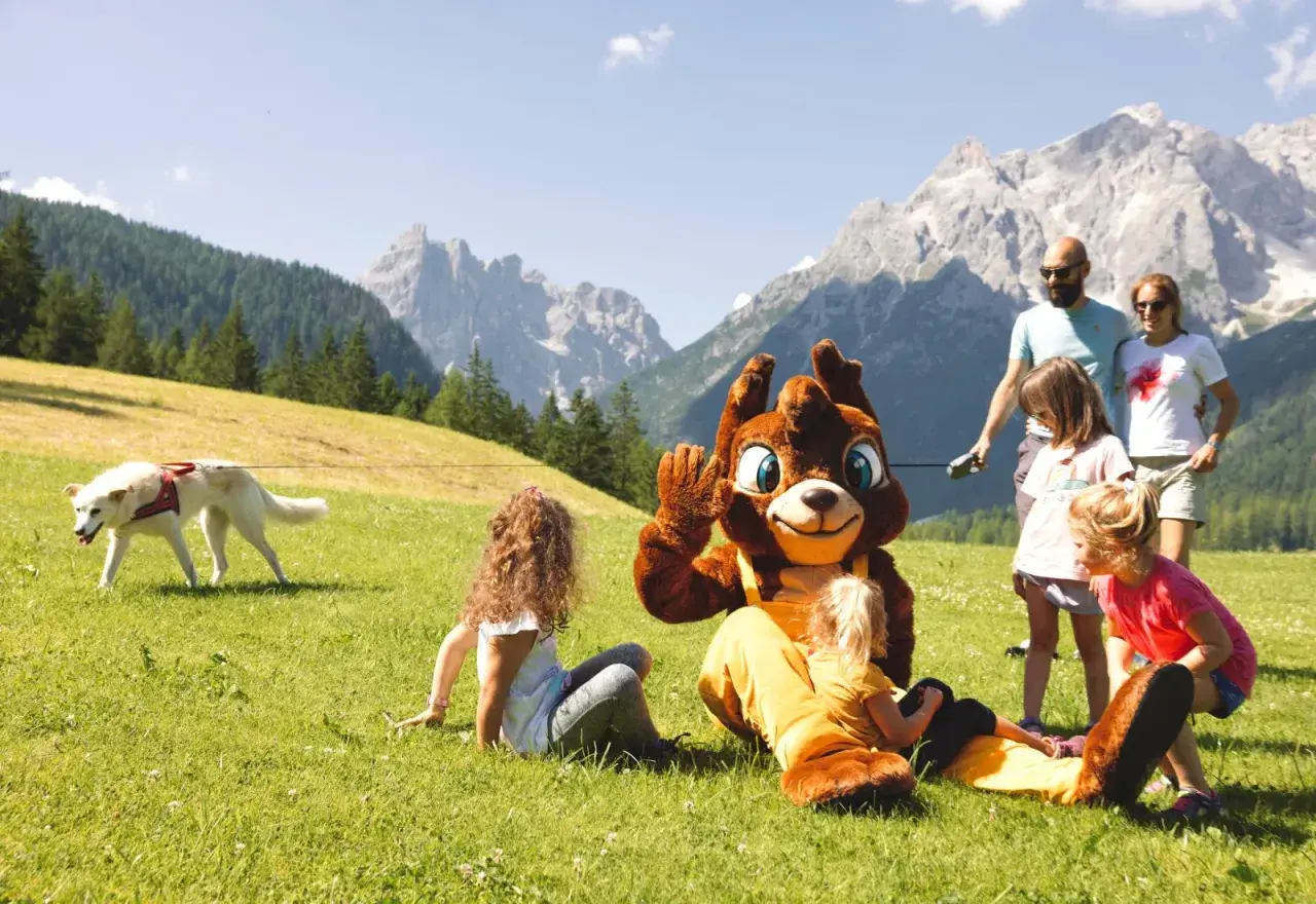 Famiglia al family resort Rainer Sesto, con mascotte e cane su prato verde, montagne sullo sfondo.