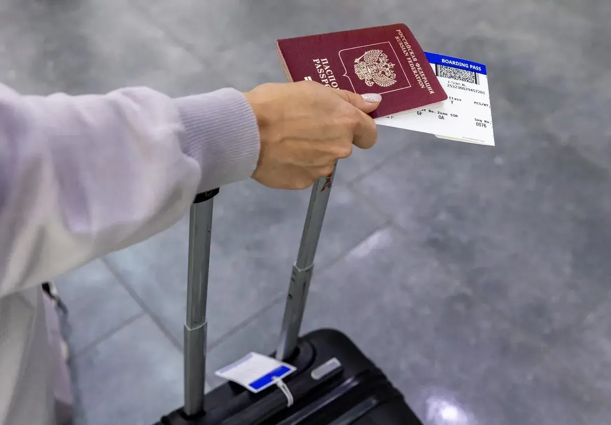 Dłoń trzyma paszport i kartę pokładową, która jest biletem na samolot. Obok walizka.