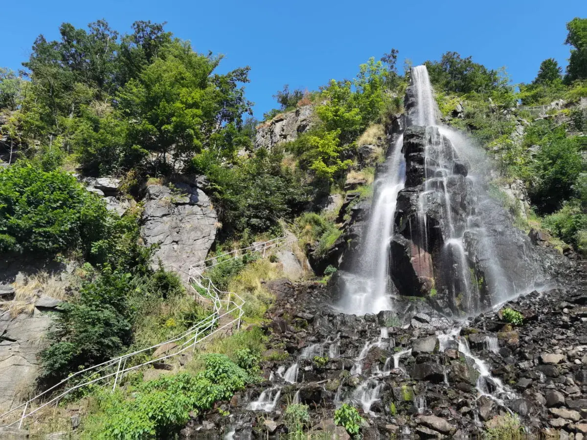 Der Trusetaler Wasserfall stürzt über schwarze Felsen in die Tiefe. Ein weißer Geländerweg führt Besucher am grünen Hang entlang.