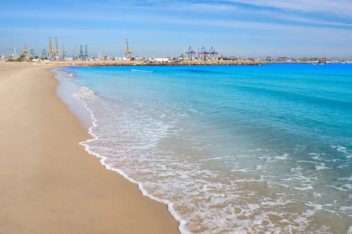 Olas suaves acarician la arena dorada en las **playas de Valencia**, con el puerto industrial de fondo bajo un cielo azul.
