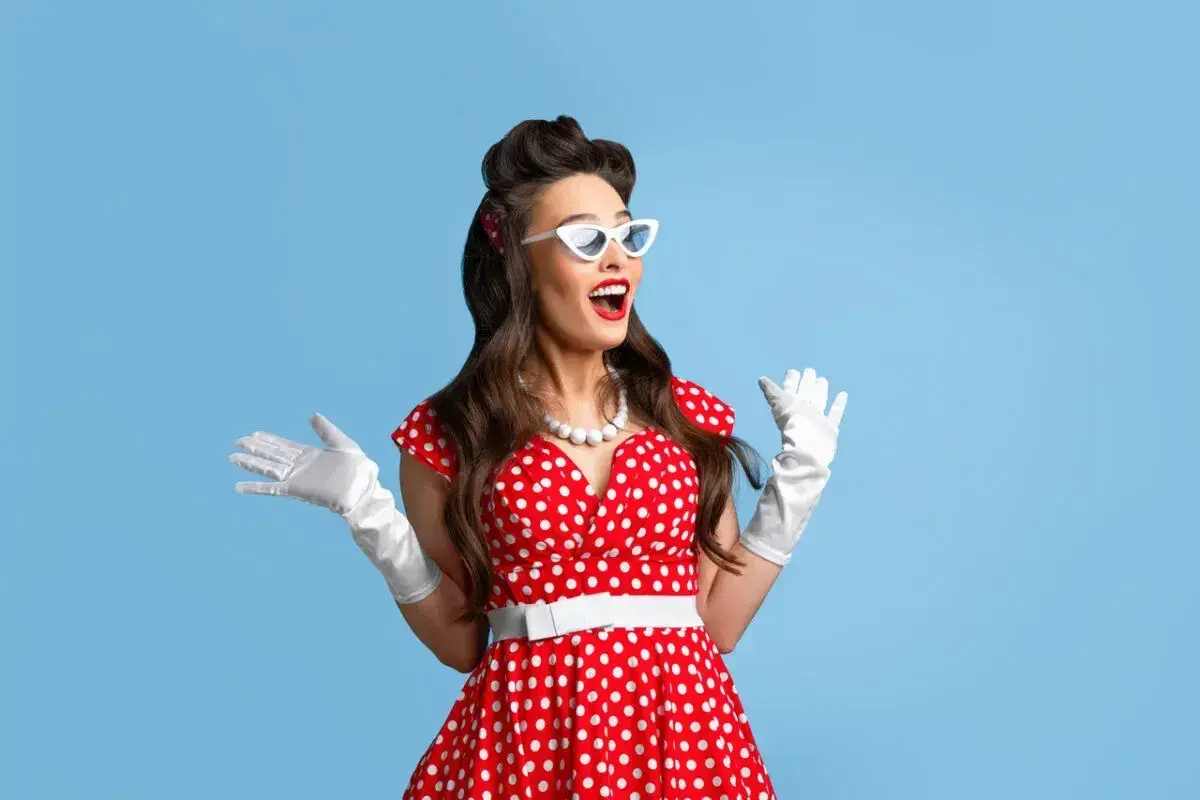 Kobieta w stylu pin-up w czerwonej sukience w białe grochy, białych rękawiczkach i okularach, z uniesionymi rękami.